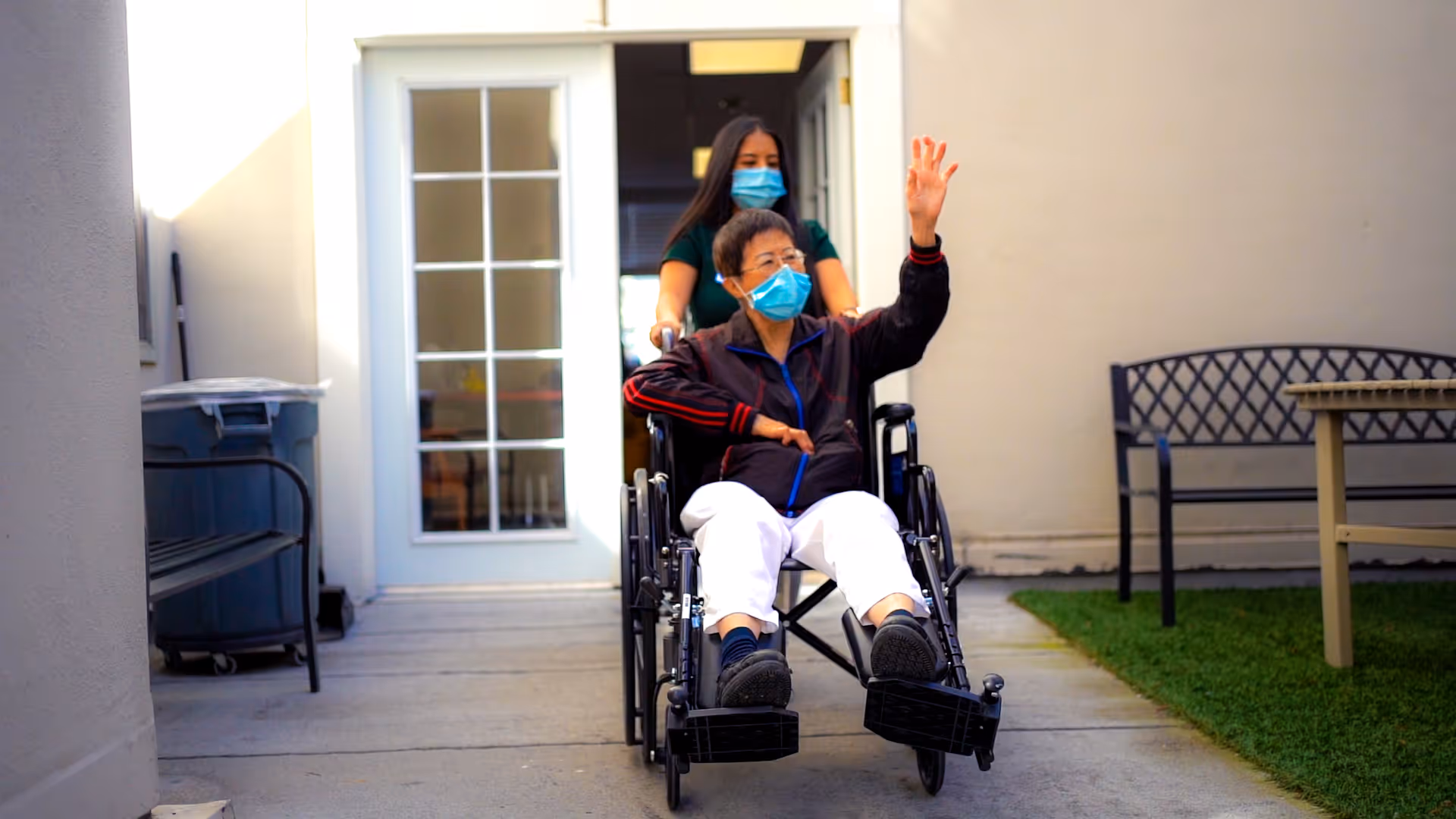 An elderly person wearing a face mask is sitting in a wheelchair being pushed by a caregiver who is also wearing a face mask. They are outside near a building entrance with a glass door, next to a bench and a trash bin.