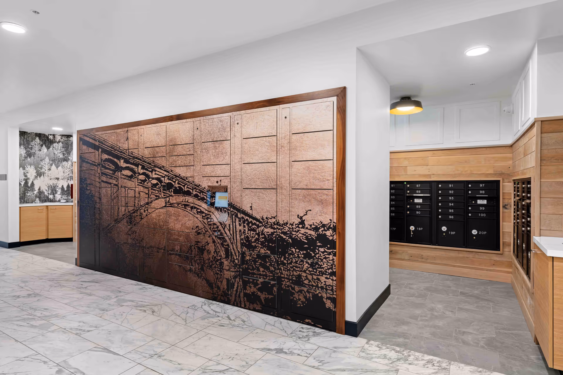 Interior view of a mailroom area in a senior living facility with a large decorative panel featuring an artistic image of a bridge on the left wall. To the right, there are multiple black mailboxes set into wooden paneling. The floor is tiled with a marble pattern, and the walls are white with wood accents. A light fixture is visible on the ceiling above the mailboxes.