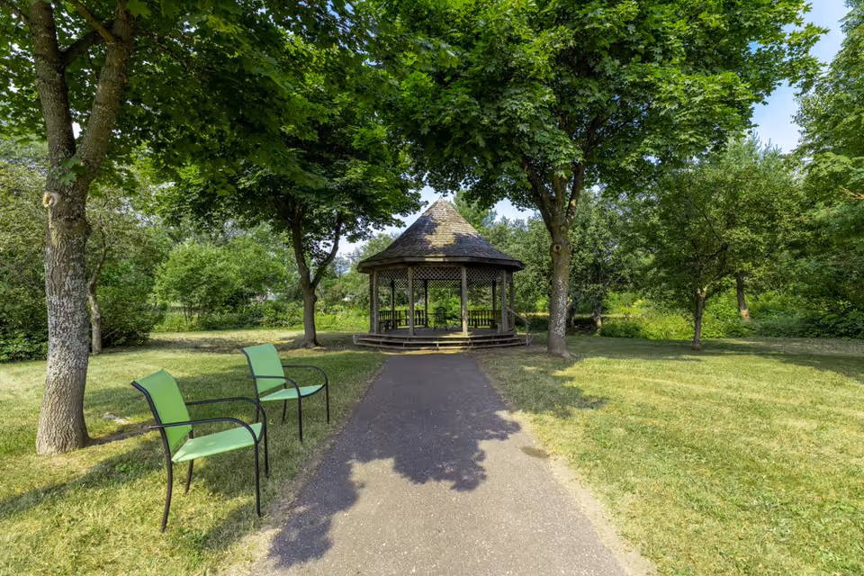 A peaceful outdoor garden area with a paved pathway leading to a wooden gazebo surrounded by green trees and grass. Two green chairs with black metal frames are placed on the left side of the pathway under the shade of the trees.