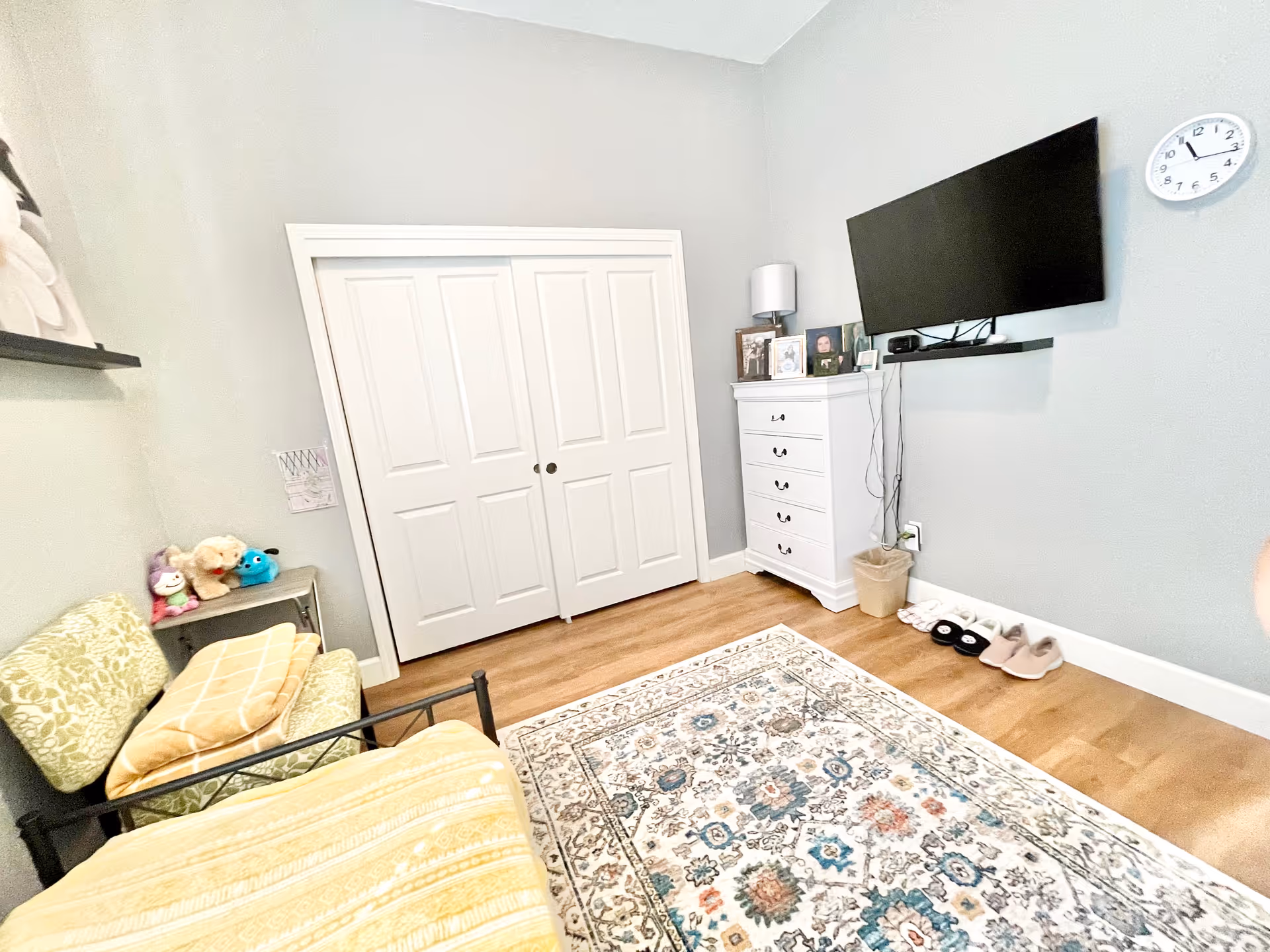 A cozy bedroom with light gray walls and wooden flooring. The room features a bed with a yellow blanket, a green patterned chair with a yellow pillow, a small side table with stuffed animals, a white dresser with framed photos and a lamp, a wall-mounted flat-screen TV, a clock on the wall, and a patterned area rug on the floor. There is a double-door closet with white doors.