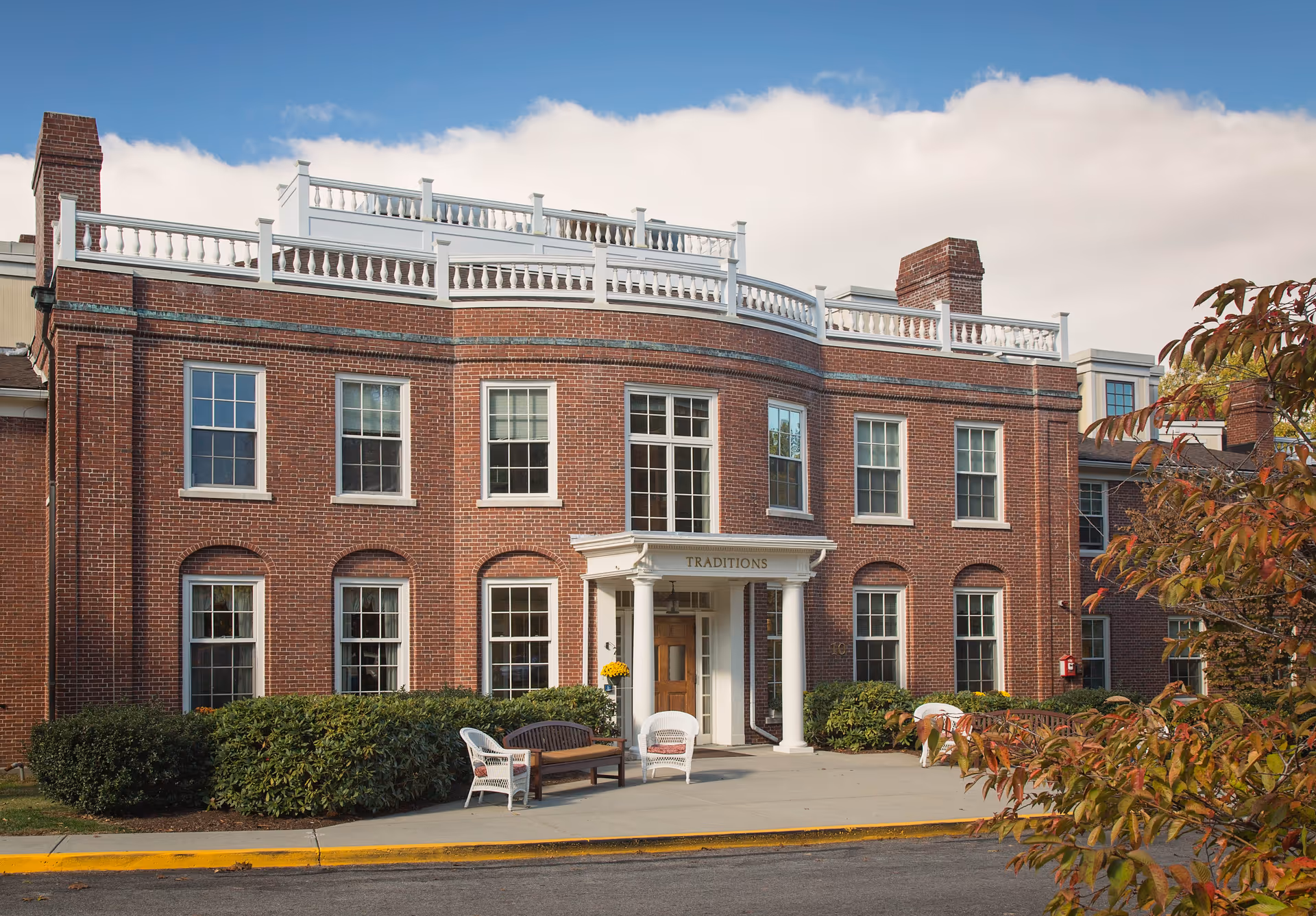 Front exterior view of a large two-story brick building with white trim and multiple windows. The entrance has a small portico with columns and the word 'TRADITIONS' above the door. There are outdoor chairs and a bench on the concrete walkway in front of the entrance, surrounded by green bushes and some autumn-colored trees.