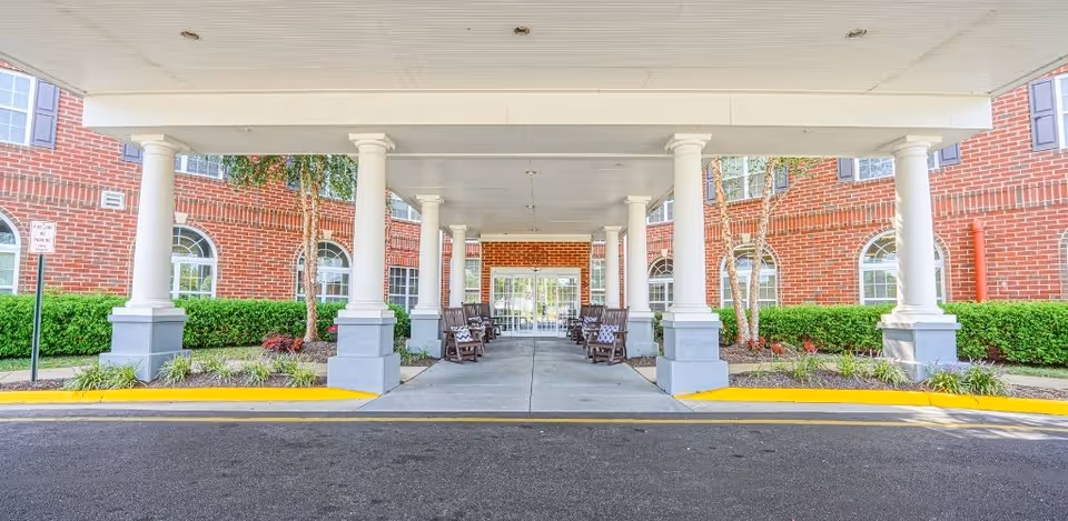 Covered drive-up entrance with white columns and benches in front of a red-brick senior living building.