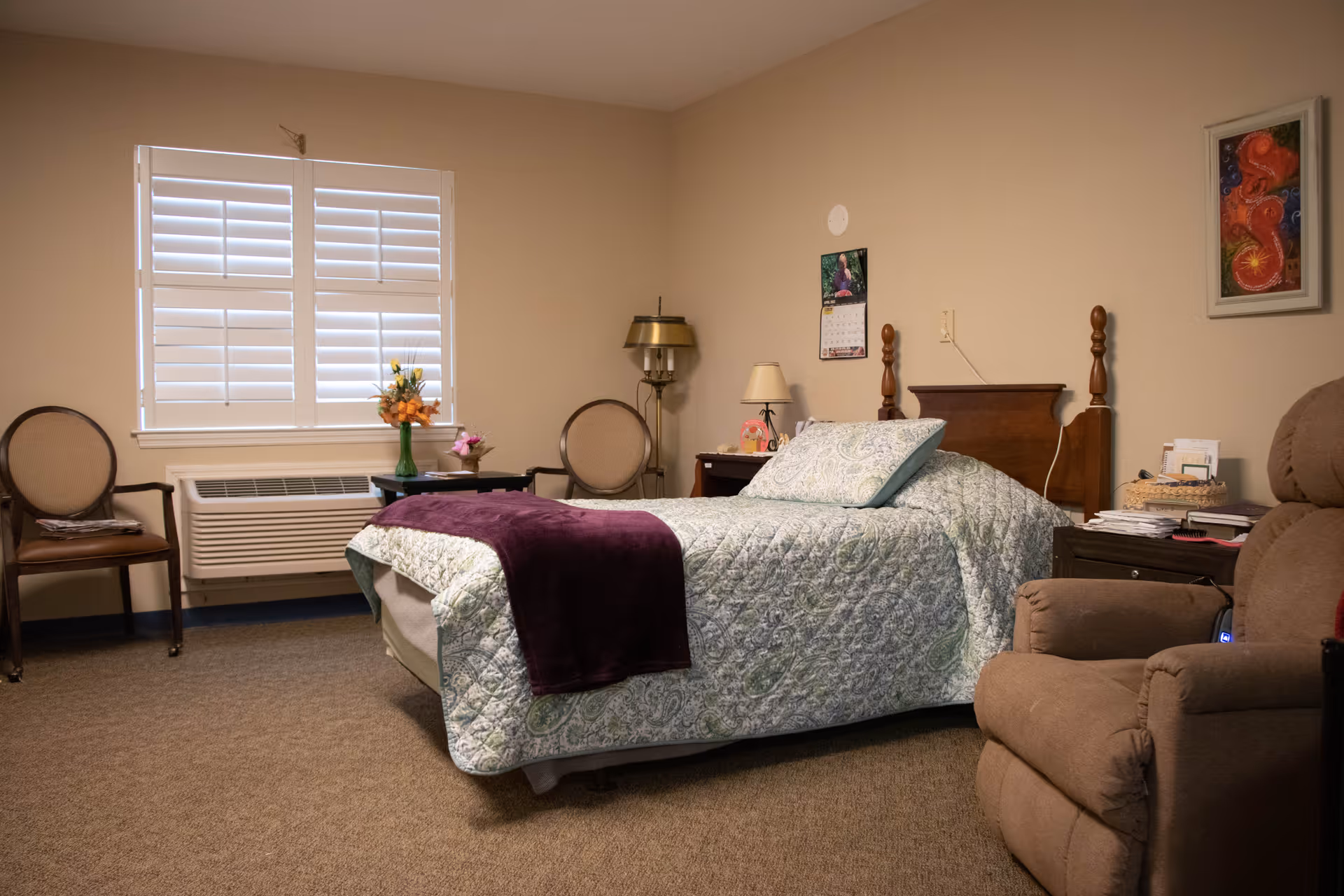 A cozy bedroom in a senior living facility featuring a single bed with a patterned quilt and a purple throw blanket. The room includes two chairs near a window with white shutters, a small table with a vase of flowers, a recliner chair, a nightstand with a lamp, and a framed picture on the wall.
