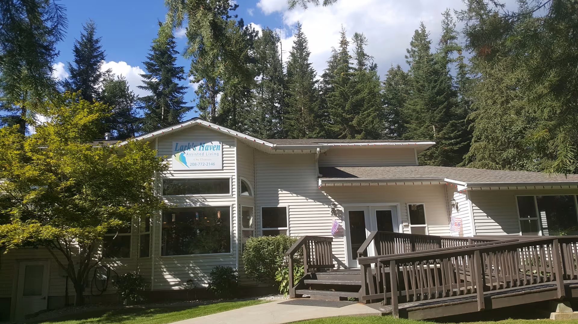 Exterior view of Larkshaven Assisted Living facility surrounded by tall pine trees and a clear blue sky. The building is light-colored with a ramp and stairs leading to the entrance. There is a sign on the building that reads 'Larkshaven Assisted Living' along with a phone number.