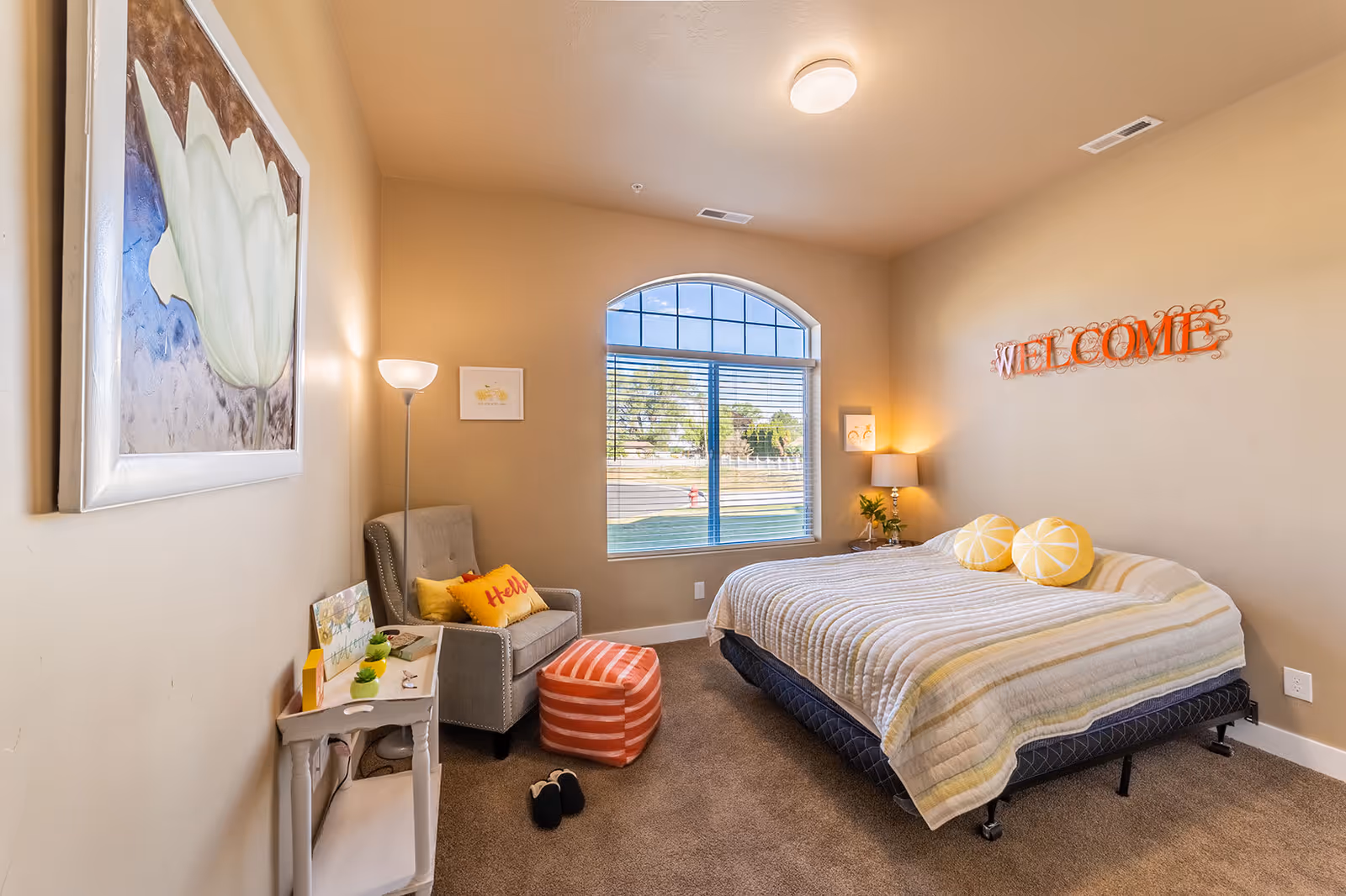 A cozy bedroom with beige walls and carpeted floor featuring a large bed with a striped quilt and two round yellow pillows. A gray armchair with yellow pillows and a striped orange ottoman are placed near a window with blinds. A small white side table holds decorative items, and a floor lamp stands beside the chair. The wall above the bed has a decorative orange 'WELCOME' sign, and a large framed flower painting hangs on the opposite wall.