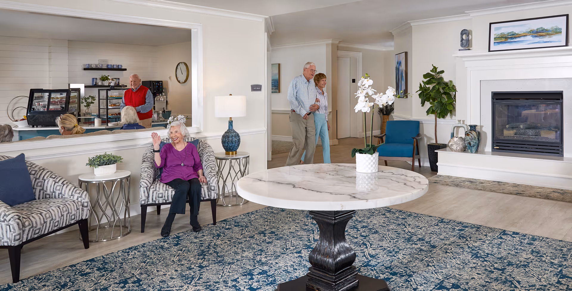 A bright and welcoming senior living facility common area with a large round marble table in the center, a blue patterned rug, and comfortable seating including patterned armchairs and a blue chair near a fireplace. An elderly woman in a purple top is sitting and waving, while an elderly couple walks in the background. A man stands behind a counter with display cases, visible through a large interior window.