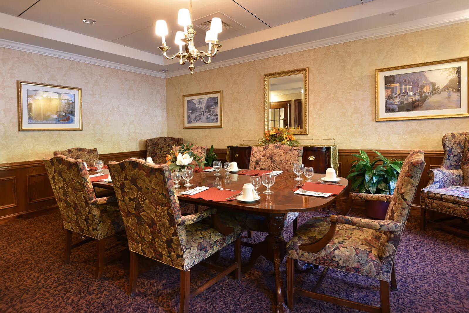 A formal dining room with a large wooden table set with glassware, cups, and red placemats. The chairs around the table have floral upholstery. The room features patterned wallpaper, framed artwork, a chandelier, a mirror, and a sideboard with a floral arrangement. There is also a potted plant and an upholstered armchair in the corner.