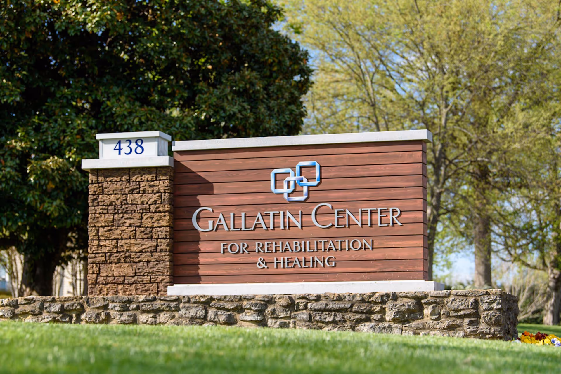 Stone and wood entrance sign reading 'Gallatin Center for Rehabilitation & Healing' with the address number 438 and trees in the background.