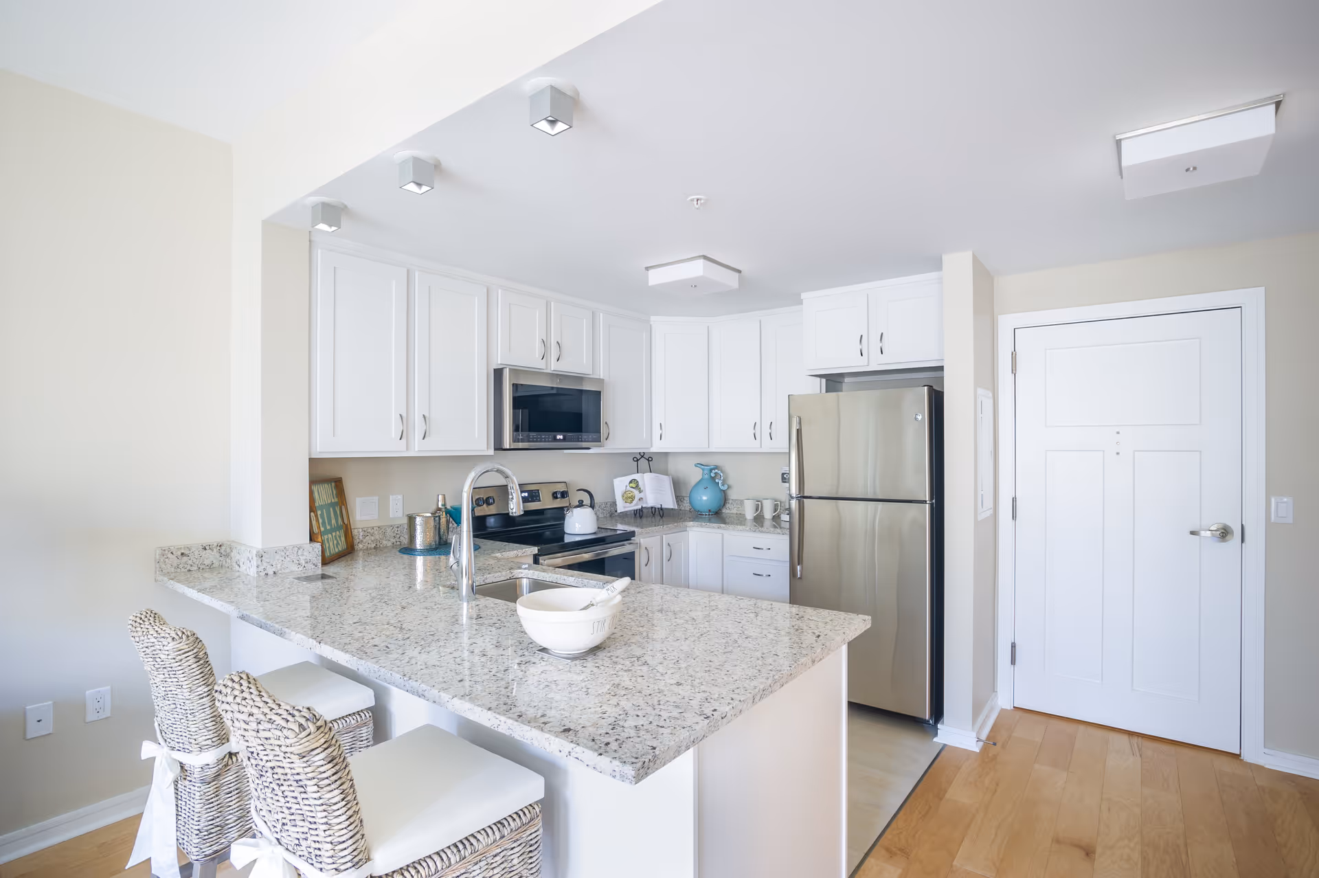 Bright modern kitchen with white cabinets, stainless steel refrigerator and stove, granite countertops, a sink with a faucet, and two wicker bar stools with white cushions at the counter.