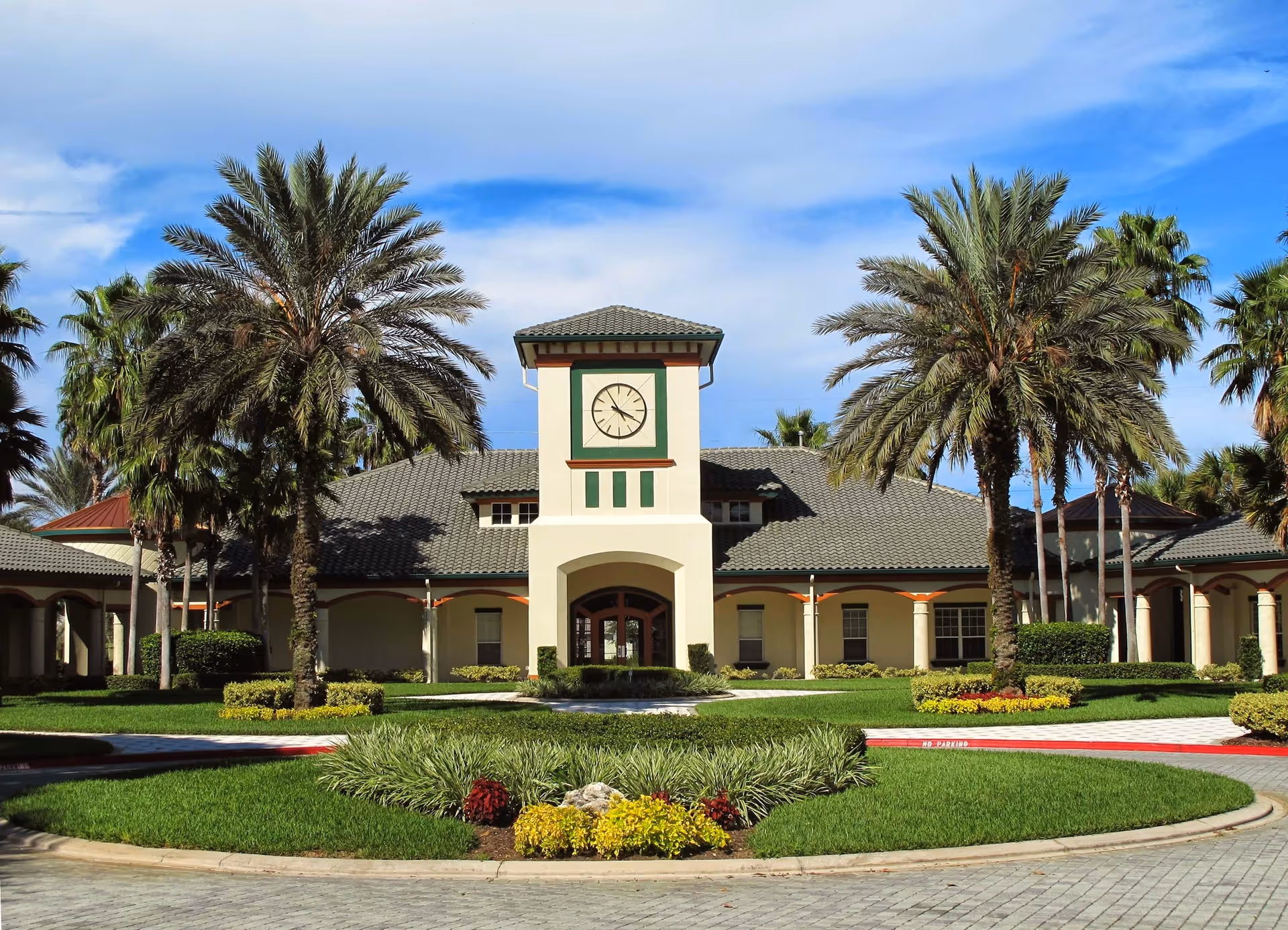 Front exterior view of a senior living facility named Legacy of Leesburg, featuring a building with a clock tower, surrounded by palm trees and well-maintained landscaping under a partly cloudy blue sky.