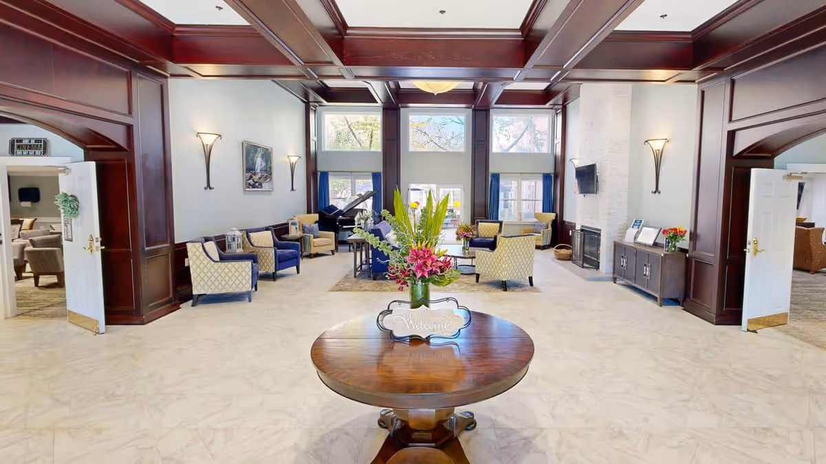 A spacious and elegant common area with high ceilings and large windows letting in natural light. The room features a round wooden table with a flower arrangement and a welcome sign in the center. Comfortable armchairs and sofas are arranged around a fireplace and a grand piano is visible near the windows. The walls have dark wood paneling and sconces providing soft lighting.