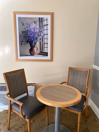 A small round wooden table with a metal pedestal base is positioned between two wooden chairs with gray cushioned seats and striped backs. Behind the table and chairs, there is a beige wall with a framed picture of purple flowers in a brown vase placed near a window.