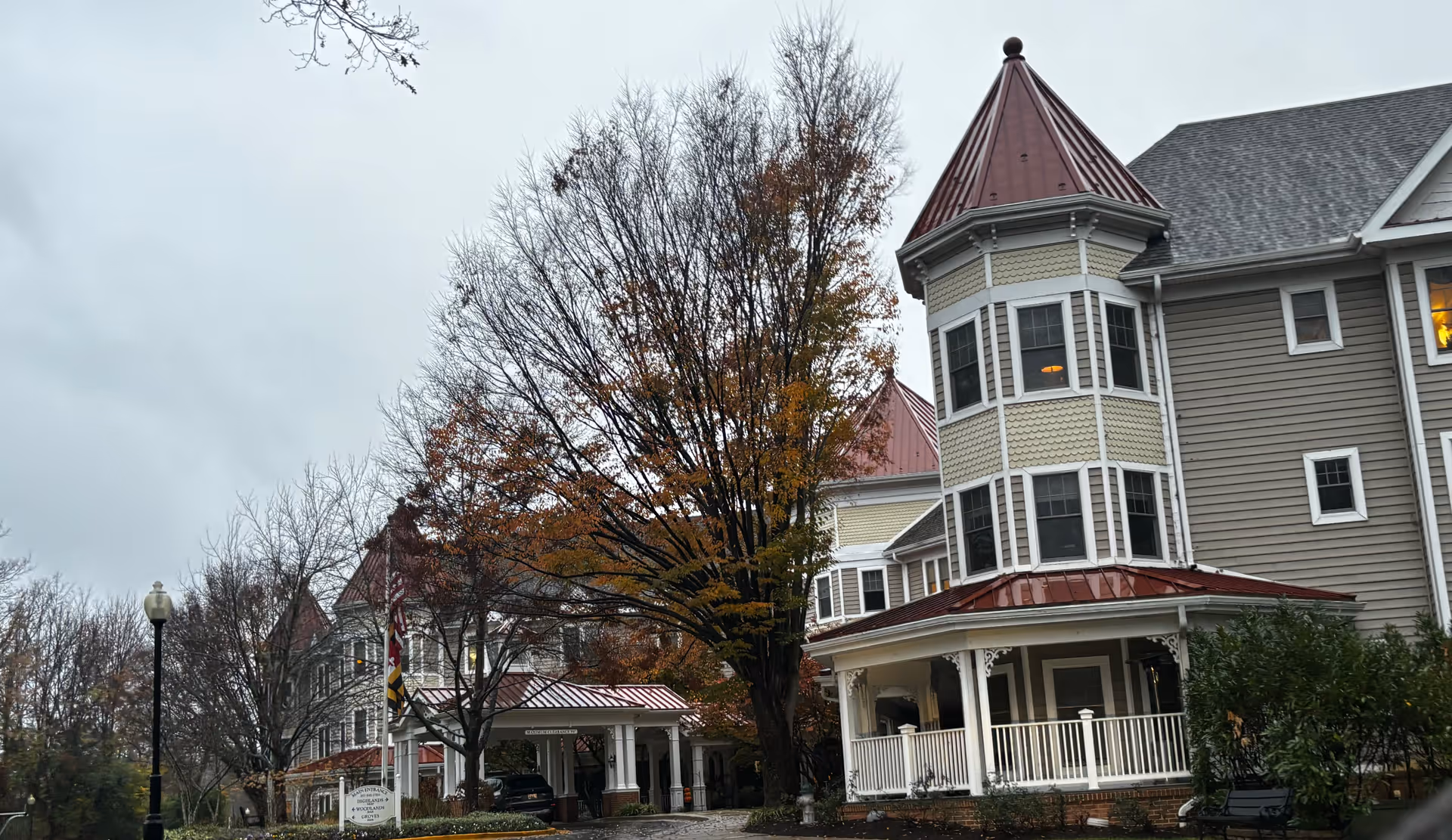 Exterior view of a senior living facility building with Victorian-style architecture, featuring a turret with a red roof, multiple windows, and a covered entrance. Trees with autumn foliage surround the building under a cloudy sky.