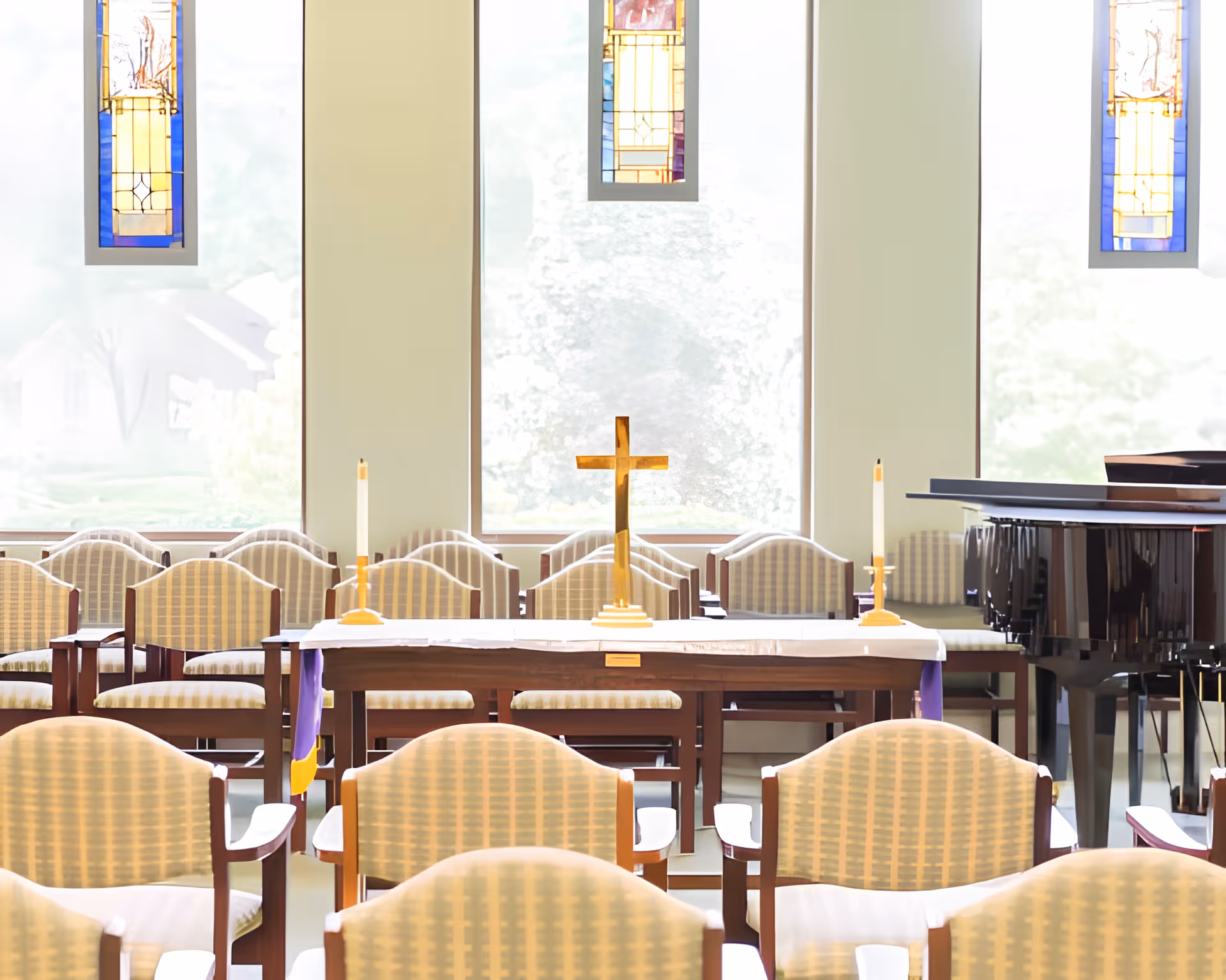 Interior view of a chapel with rows of cushioned chairs facing a wooden altar table. On the altar are two lit candles and a wooden cross. Behind the altar are large windows with stained glass panels, allowing natural light to fill the space. A grand piano is visible to the right side of the altar.
