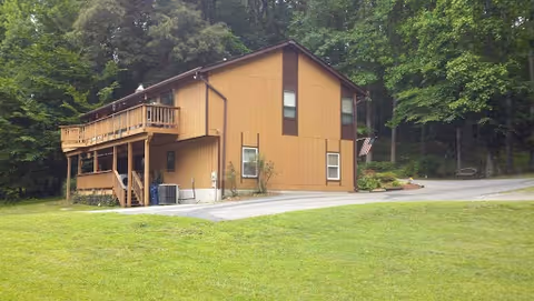 A two-story brown building with a wooden balcony on the upper floor, surrounded by green grass and trees in the background.