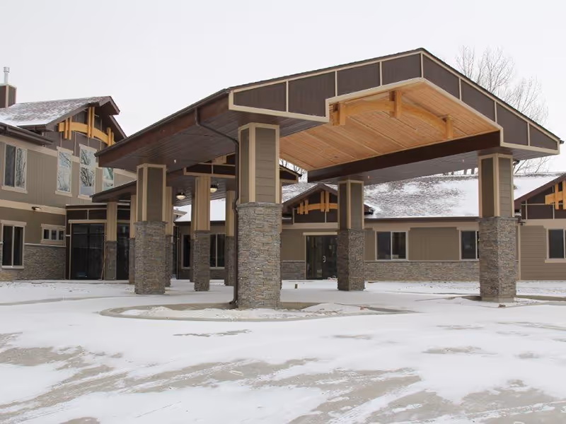 Front entrance of a lodge-style building with a large covered porte-cochere supported by stone columns and snow on the ground.