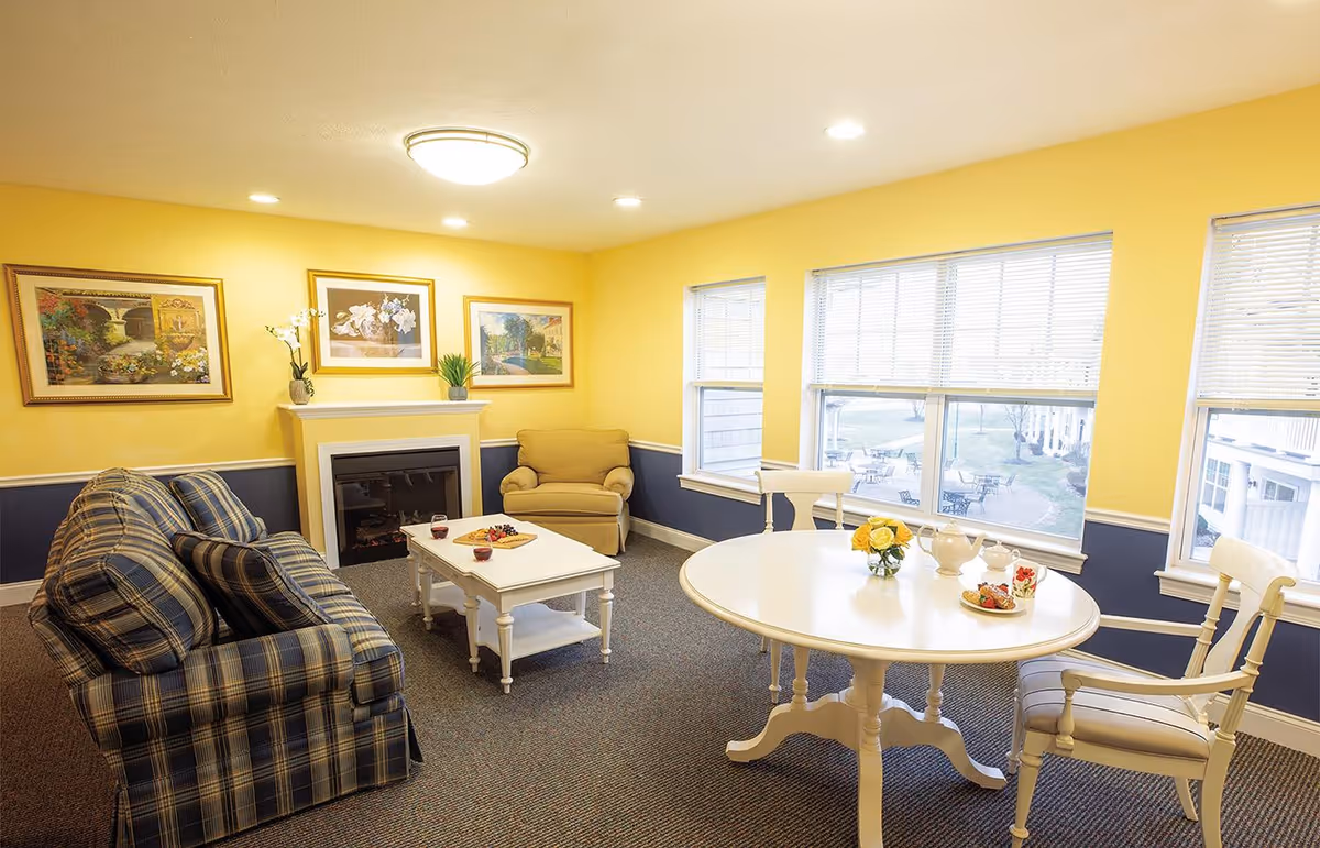 Well-lit senior living common room with a plaid sofa, armchair, fireplace, coffee table, and a round dining table by large windows.