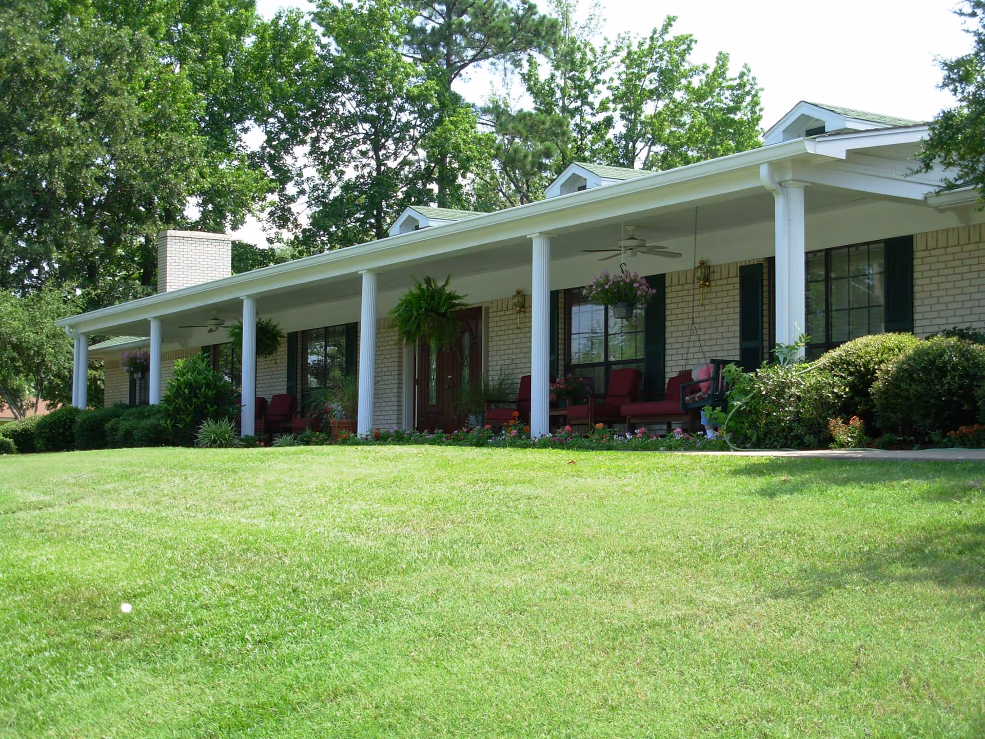 A single-story brick building with a long covered porch supported by white columns. The porch has hanging plants, ceiling fans, and red cushioned outdoor furniture. The building is surrounded by green grass and bushes, with trees in the background under a clear sky.