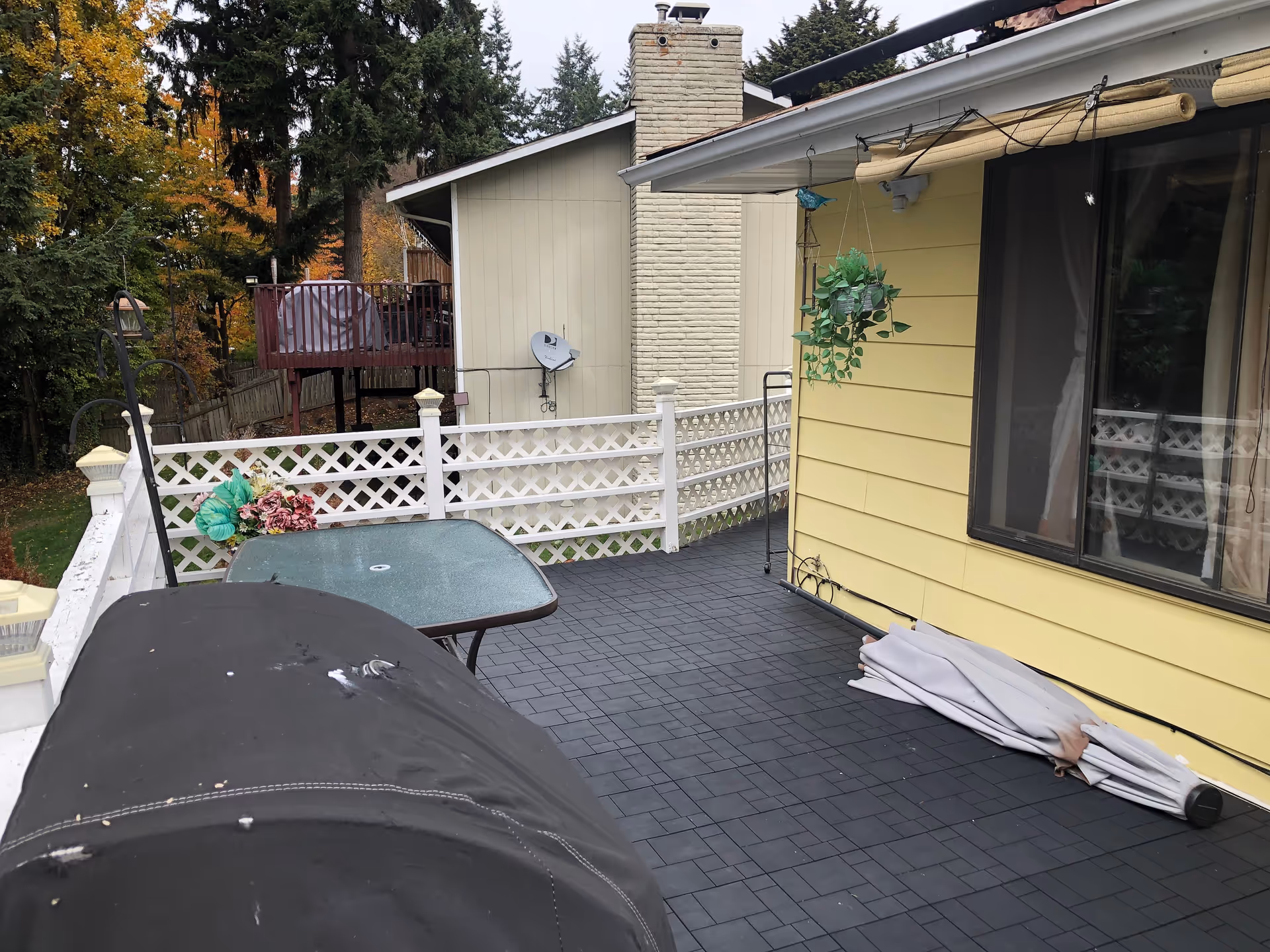 Outdoor patio area with a black grill covered by a black cover, a glass-top table with a bouquet of artificial flowers, white lattice fencing, a yellow house wall with a window, a hanging plant, and a folded umbrella on the floor. Trees and another building are visible in the background.