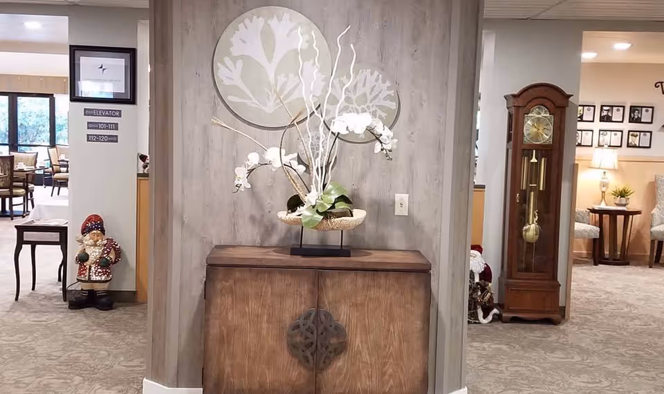 Interior view of a senior living facility hallway with a wooden cabinet topped with a decorative floral arrangement in the center. To the left, there is a small statue of a person in winter clothing and signs indicating elevator directions. To the right, there is a tall grandfather clock and a small table with a lamp and plant, with framed pictures on the wall in the background.