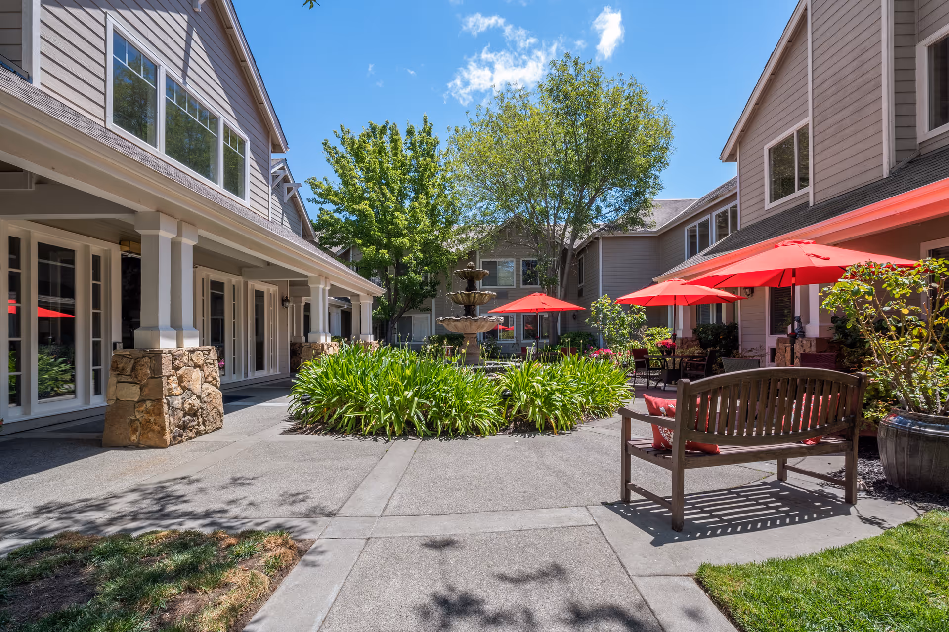 Outdoor courtyard area of Aegis Living Corte Madera featuring a central three-tiered stone fountain surrounded by green plants. The courtyard is flanked by two beige buildings with large windows and stone pillars. There are red umbrellas shading tables and chairs on the right side, and a wooden bench with red cushions in the foreground. Trees and a clear blue sky are visible in the background.