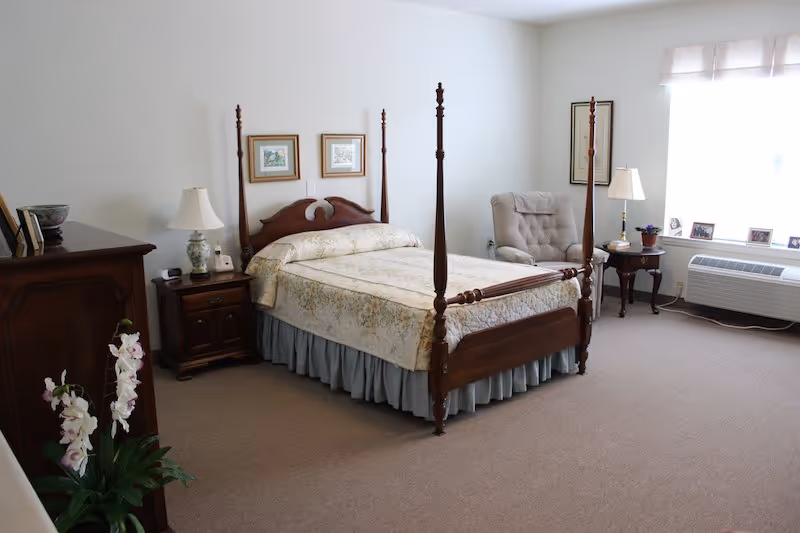A bedroom with a wooden four-poster bed covered with a floral bedspread. There is a wooden nightstand with a lamp and a clock beside the bed. A cushioned armchair is placed near a window with blinds, next to a small wooden side table with a lamp and framed photos. A wooden dresser and a potted plant with white flowers are also visible in the room.