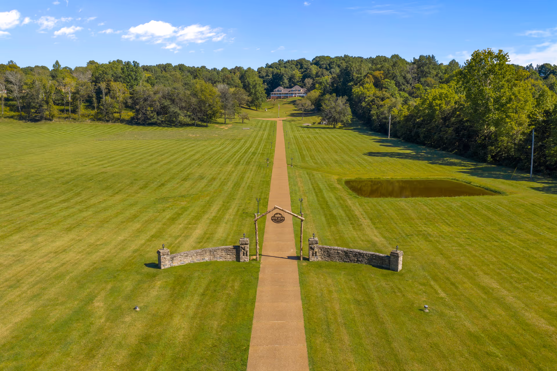 A long paved walkway leads through a large, well-maintained grassy field towards a building in the distance surrounded by trees. The entrance to the walkway features a stone wall with pillars and a wooden arch with a sign that reads 'New Hope'. There is a small pond on the right side of the field and dense trees border the area under a blue sky with scattered clouds.