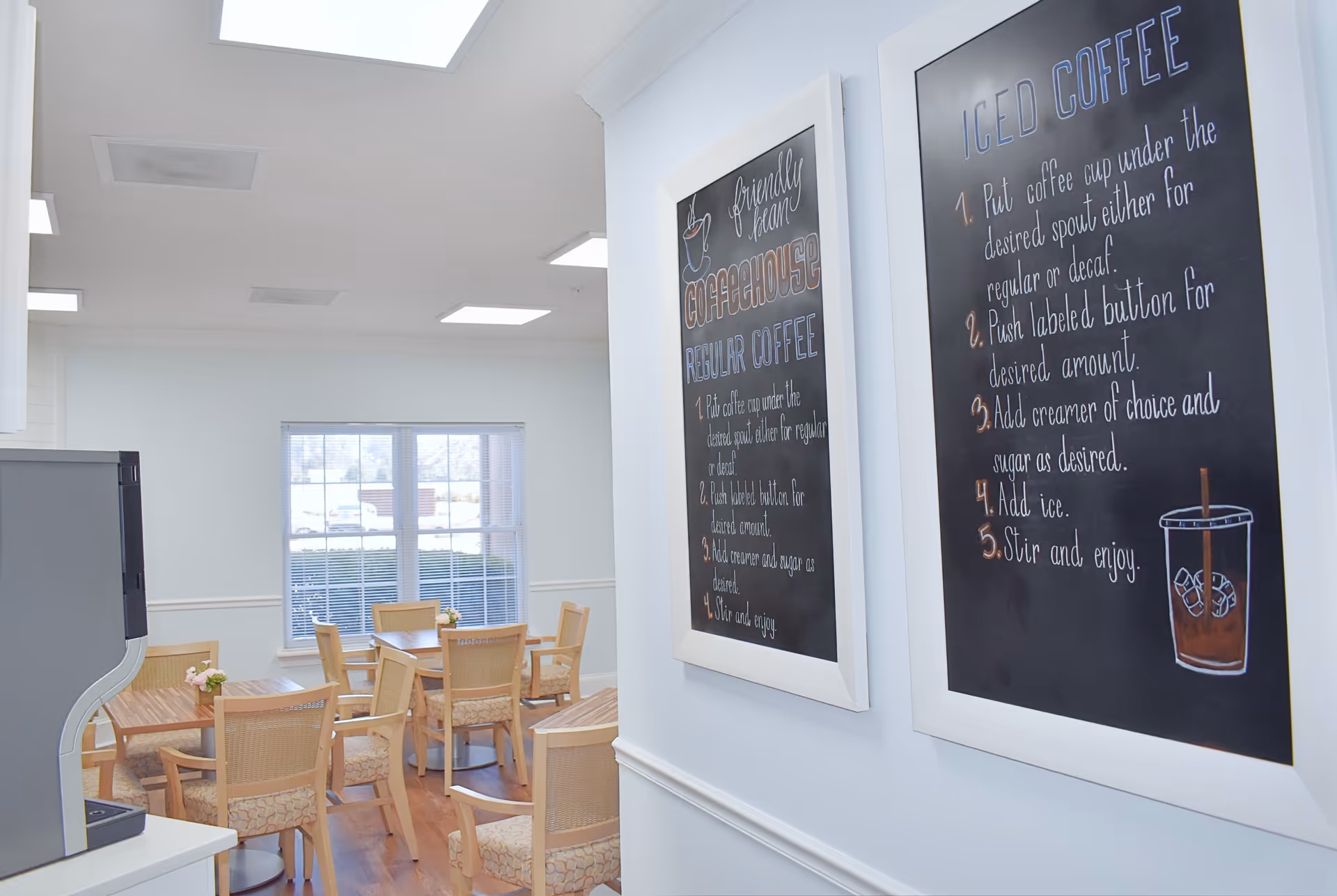 Interior view of a bright coffeehouse area with wooden tables and chairs arranged neatly. Two framed chalkboard-style signs on the wall provide instructions for making regular coffee and iced coffee. A large window lets in natural light, and a coffee machine is partially visible on the left side.
