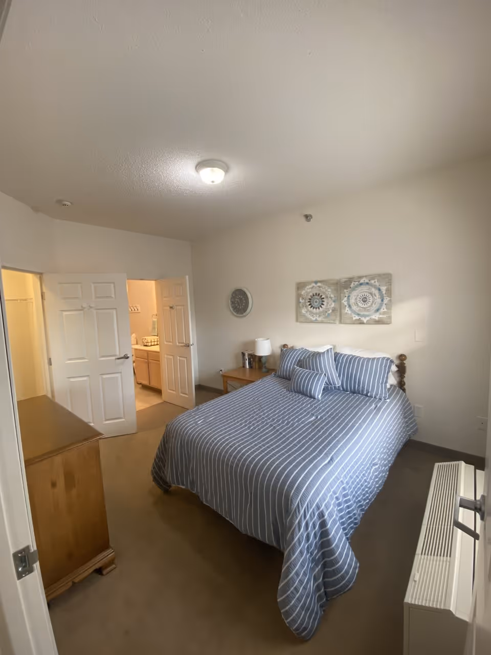 A bedroom with a bed covered in blue and white striped bedding, two decorative pillows, and a wooden headboard. There is a wooden dresser on the left side and a nightstand with a lamp and books on the right side. The walls are light-colored with two decorative wall hangings above the bed and a round clock on the adjacent wall. An open door reveals a bathroom and a closet area.