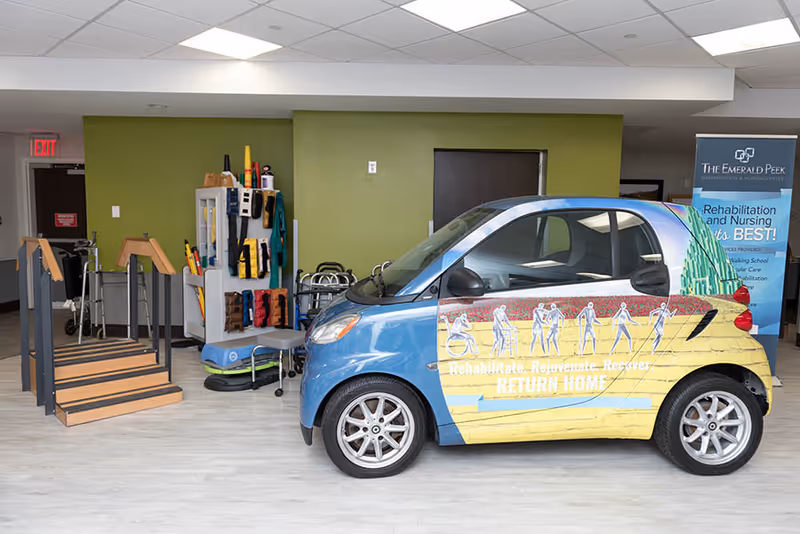 Indoor rehabilitation area with a small blue and yellow car featuring illustrations of people using mobility aids and the words 'Rehabilitate. Rejuvenate. Recover. RETURN HOME.' There is a wooden ramp, various therapy equipment, and a banner for The Emerald Peek Rehabilitation & Nursing Center in the background.