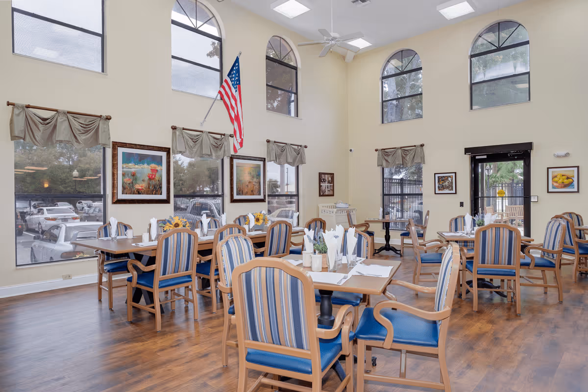 A bright dining room with multiple wooden tables and blue-striped cushioned chairs. The room features large arched windows with beige curtains, framed artwork on the walls, and an American flag hanging near the windows. The tables are set with napkins, cups, and small floral centerpieces. The floor is wooden, and there is a glass door leading outside.