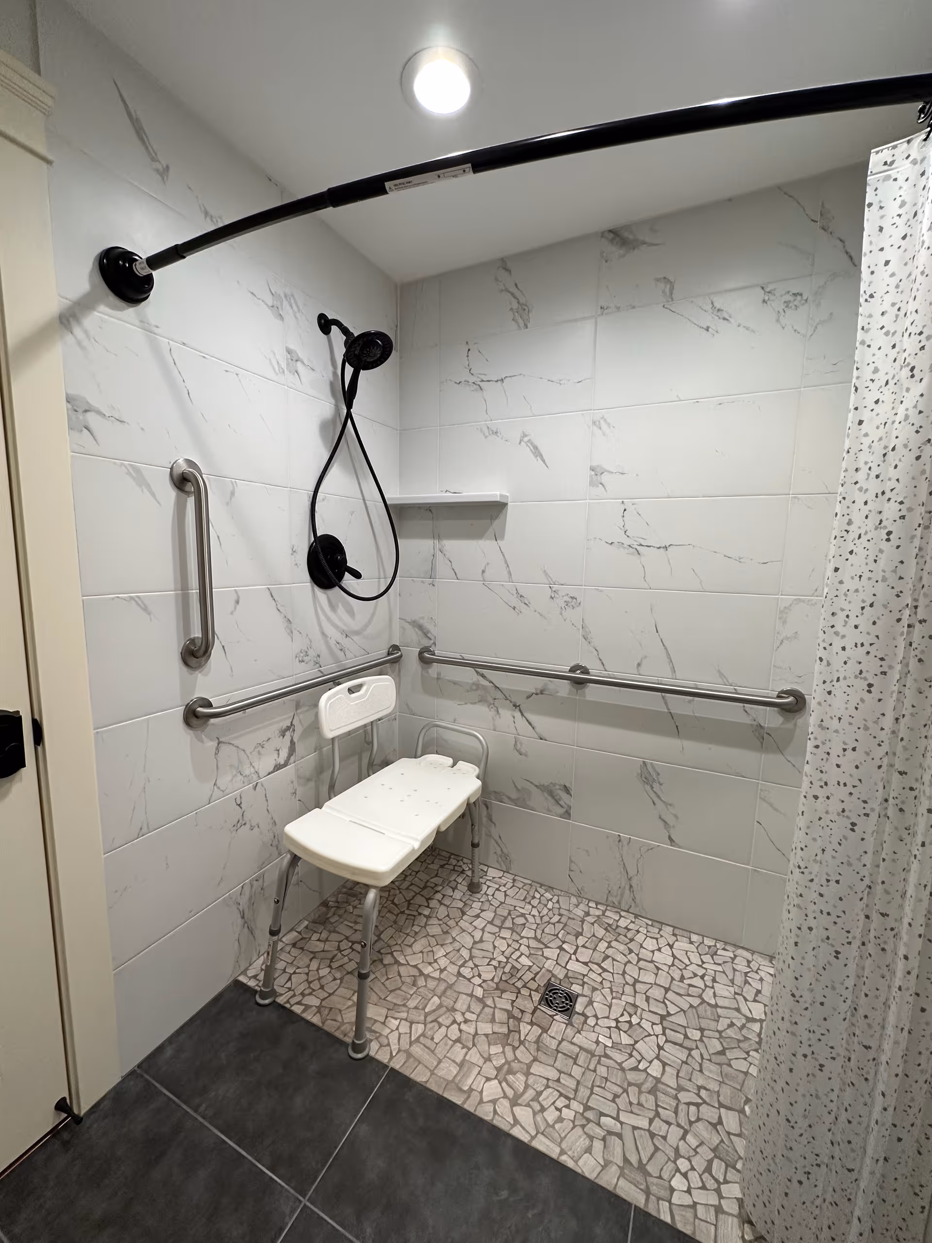 Accessible shower area with white marble-patterned tiles on the walls and mosaic tile flooring. The shower features a black handheld showerhead, a white shower chair with backrest, and multiple stainless steel grab bars on the walls. A curved shower curtain rod with a white curtain with small patterns is partially visible.