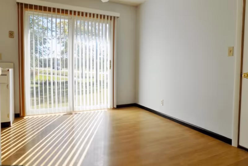 Empty room with light wood flooring and white walls, featuring a large sliding glass door with vertical blinds partially open, allowing sunlight to cast striped shadows on the floor. A small portion of a kitchen counter is visible on the left side.
