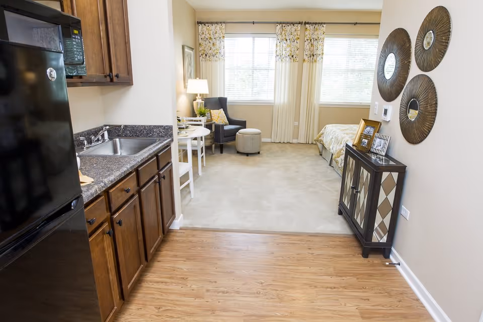 View of a studio apartment in an assisted living facility showing a small kitchen area with dark wood cabinets, a black refrigerator, and a sink on the left. The room extends into a living and sleeping area with a small round dining table with two chairs, a cushioned armchair with an ottoman, a bed with patterned bedding, and large windows with floral curtains. On the right wall, there is a decorative cabinet with framed photos and three round wall mirrors above it.