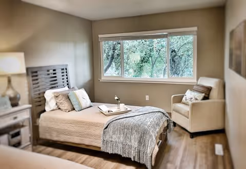A cozy bedroom with a single bed made with beige bedding, decorative pillows, and a gray throw blanket. A tray with a flower and a book is placed on the bed. Next to the bed is a nightstand with a lamp. There is a beige armchair with a pillow near a large window showing green trees outside. The room has wooden flooring and neutral-colored walls.