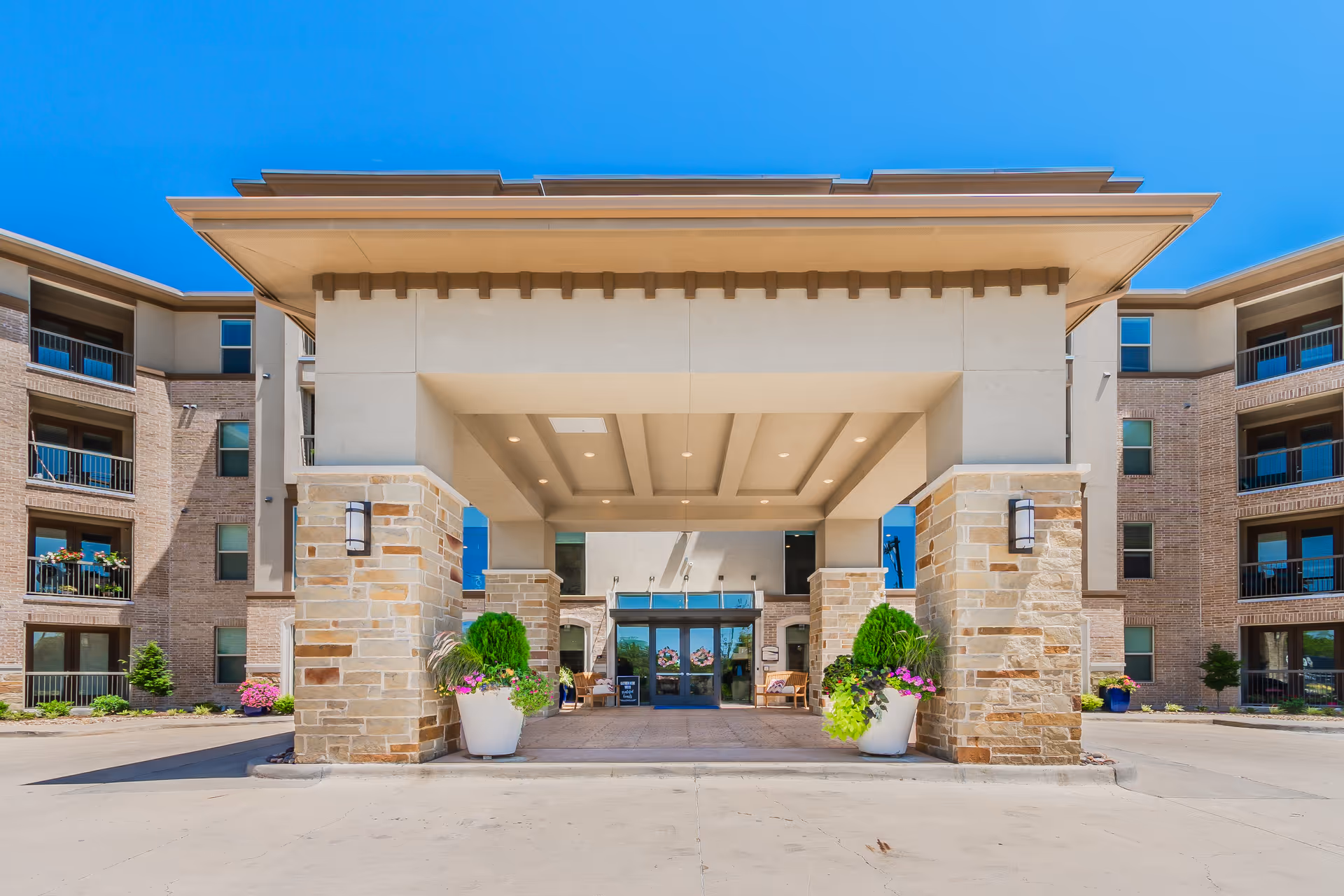 Front entrance of a multi-story residential building with a covered porte-cochere, stone columns, potted plants, and balconies.