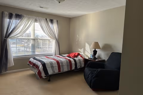 A bedroom with a single bed covered with a striped red, white, and black blanket, positioned near a large window with sheer white and dark curtains. Next to the bed is a small wooden nightstand with a lamp and a box of tissues. There is a black armchair beside the nightstand. The room has beige walls and carpeted floor.