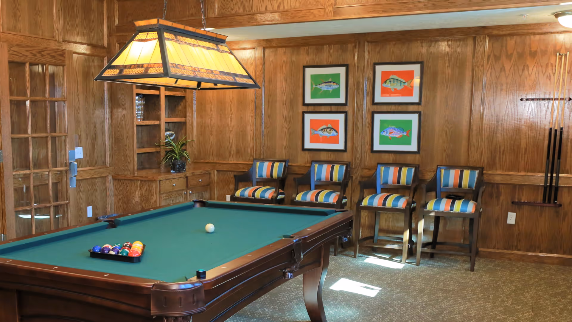 A wood-paneled game room with a pool table, hanging light fixture, striped chairs, and framed fish artwork on the wall.