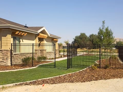 Single-story building with stone accents beside a fenced grassy courtyard and pathway under a clear blue sky.