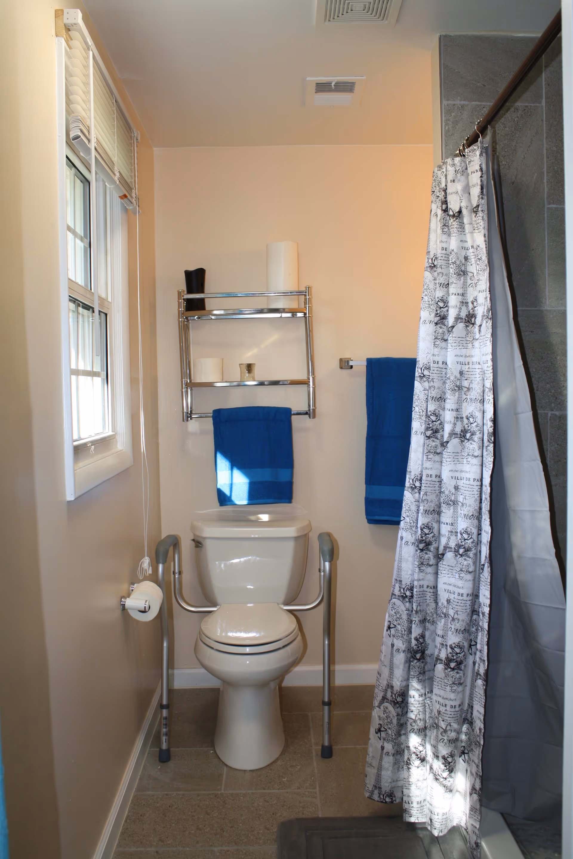 A bathroom with a white toilet equipped with safety rails on both sides. Above the toilet is a metal shelf holding a black container, a roll of paper towels, two rolls of toilet paper, and a small cup. Two blue towels hang on the wall, one on the shelf and one on a towel rack. To the right is a shower with a curtain featuring a black and white pattern. A window with white blinds is on the left wall.
