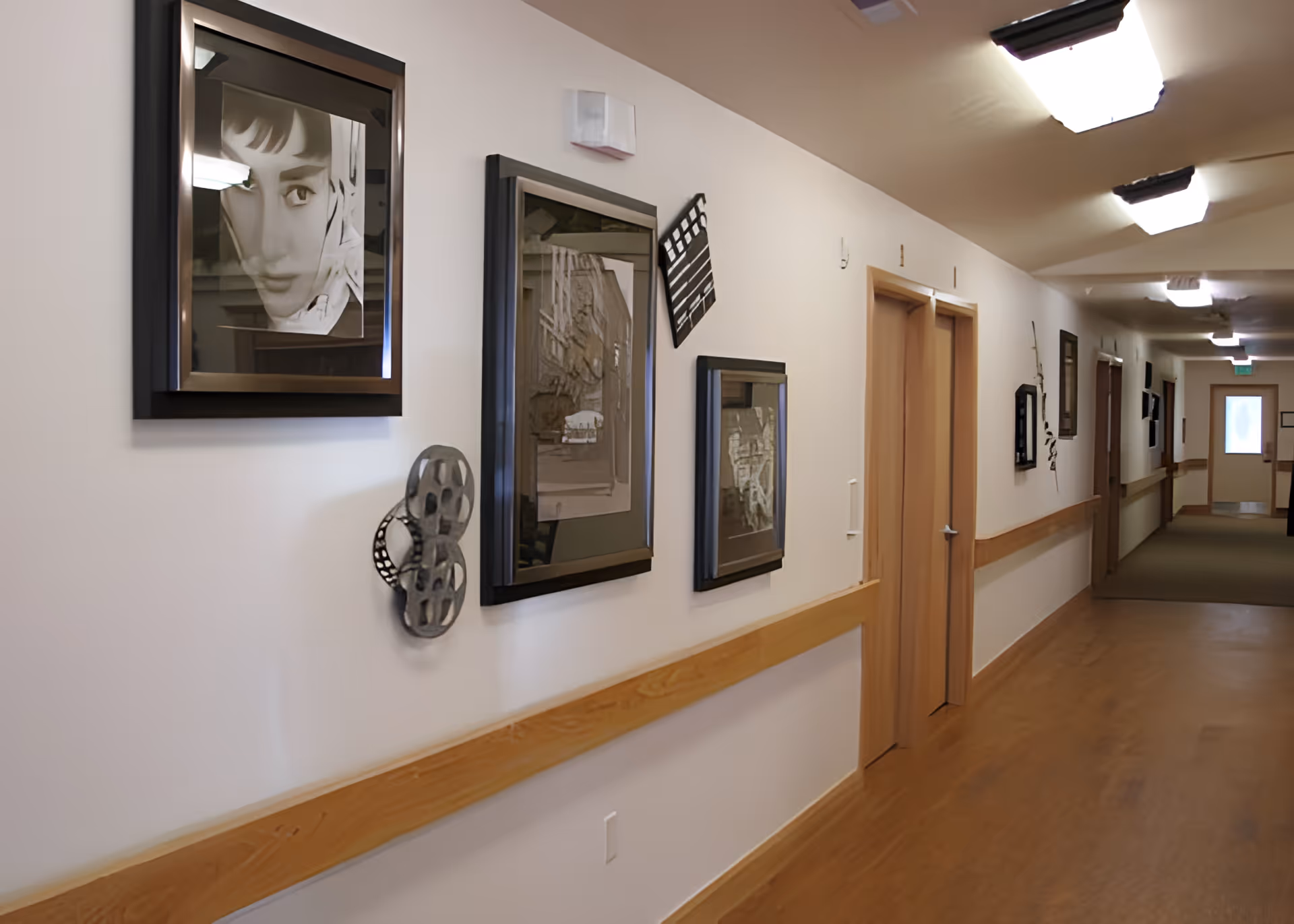 A long hallway with wooden flooring and light-colored walls decorated with framed black and white photographs and film-themed wall art, including a film reel and clapperboard. Several closed wooden doors line the hallway, and ceiling lights illuminate the space.