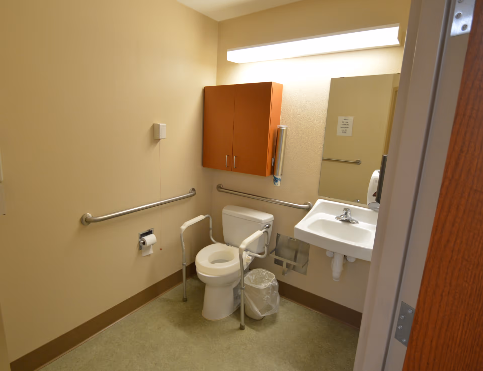 A small accessible bathroom with beige walls, a white toilet equipped with safety rails, a white sink with a mirror above it, a wooden cabinet mounted on the wall, a trash can, and stainless steel grab bars along the walls.