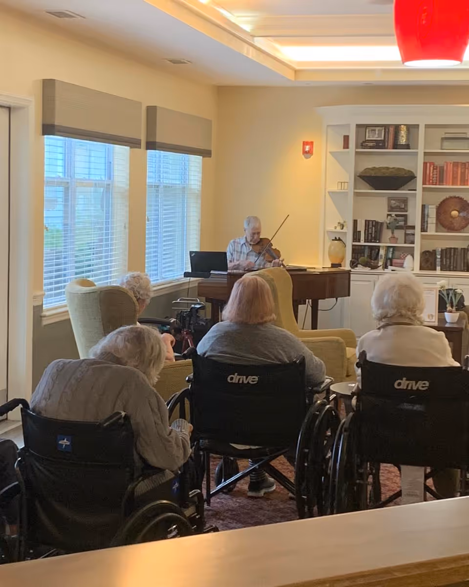 A group of elderly individuals, some in wheelchairs, seated in a living room area facing a man playing the violin near a window. The room has large windows with blinds, a bookshelf filled with books and decorative items, and comfortable chairs.