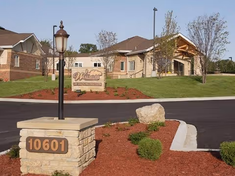 Exterior view of Wealshire of Bloomington senior living facility showing a stone sign with the address 10601 in the foreground, a lamp post, landscaped bushes and mulch, and the building with a covered entrance in the background under a clear sky.