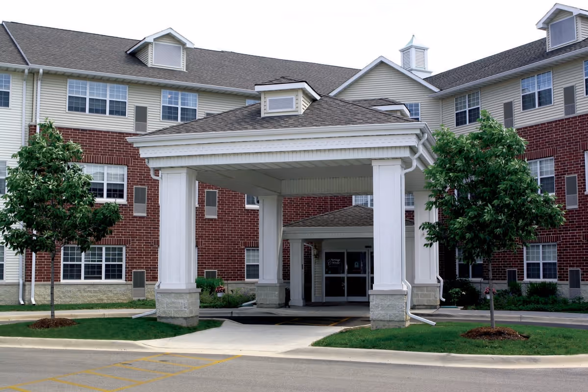 Entrance of a senior living facility with a covered drop-off area supported by white columns. The building has a combination of red brick and beige siding with multiple windows. There are two small trees planted on either side of the entrance driveway.
