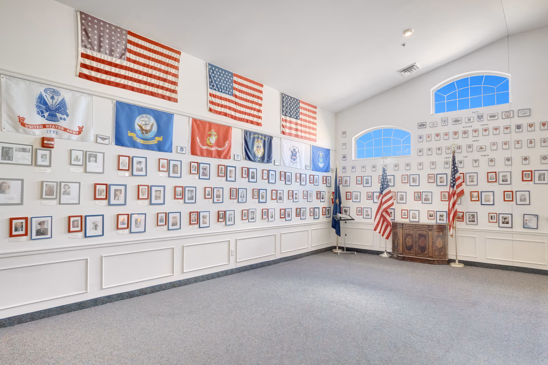 A large, bright room with white walls displaying numerous framed photographs and military flags, including several American flags and flags representing different branches of the United States military. The room has high ceilings with arched windows letting in natural light, and two American flags on stands near a wooden cabinet in the corner.