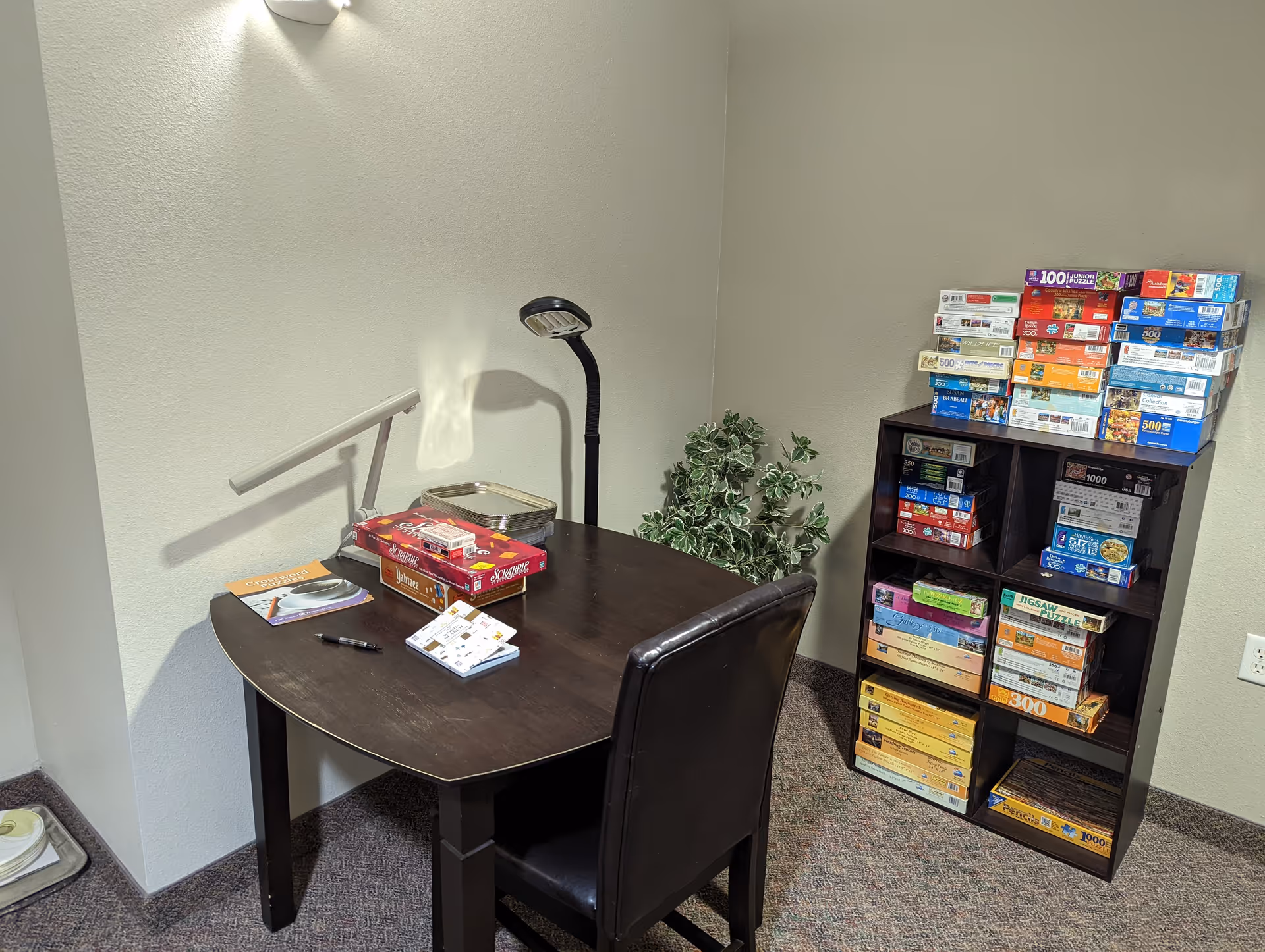 A small room corner with a dark wooden table and a black chair. On the table are board games, a pen, a small notebook, and a desk lamp. Next to the table is a small plant, and against the wall is a black shelving unit filled with various board games and puzzles. The room has beige walls and carpeted floor.