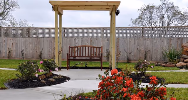 A wooden bench under a wooden pergola in a garden area with a concrete pathway, surrounded by green grass, flowering bushes with red and pink flowers, and a wooden fence in the background.