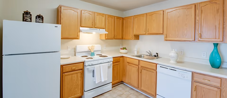 Bright L-shaped kitchen with oak cabinets, white refrigerator, stove, dishwasher, and a sink on a light countertop.
