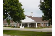 Front exterior of a single-story brick healthcare building with a covered entrance, cupola, bench, and surrounding trees.